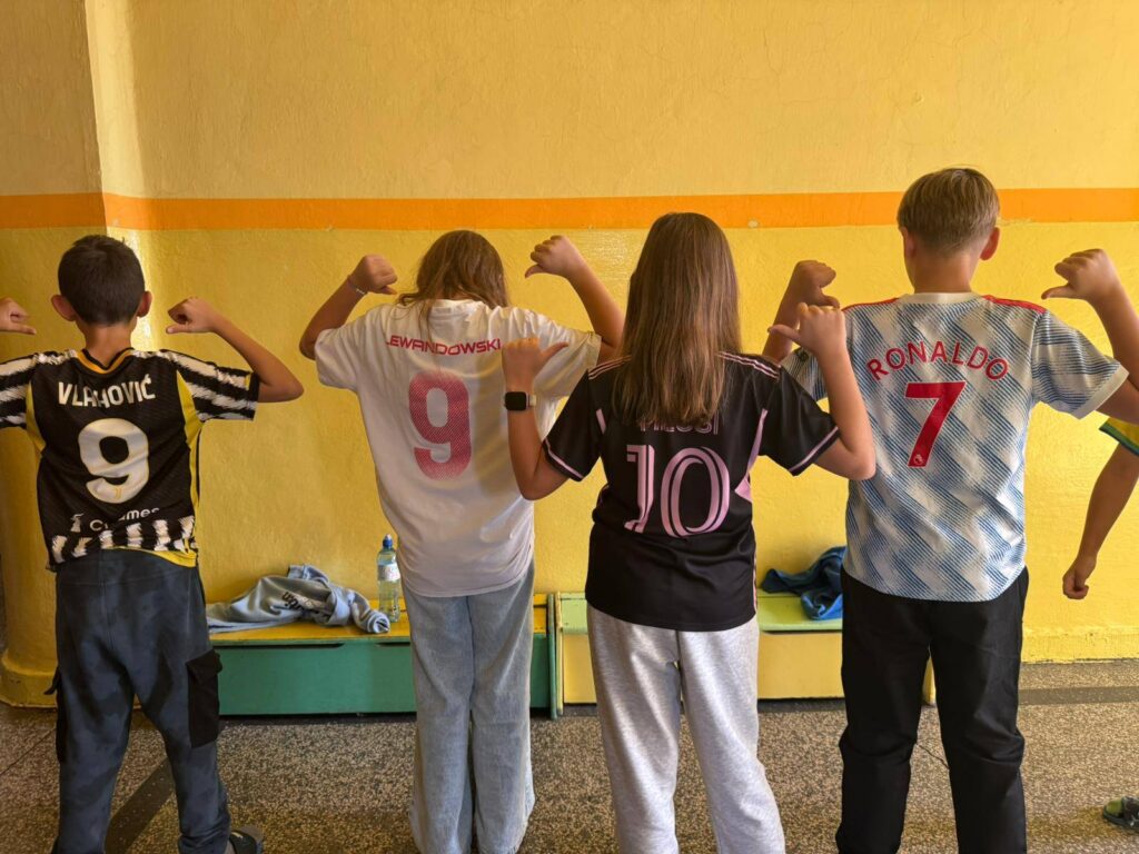 Uczniowie stoją plecami na szkolnym korytarzu, prezentując różne koszulki drużyn piłkarskich