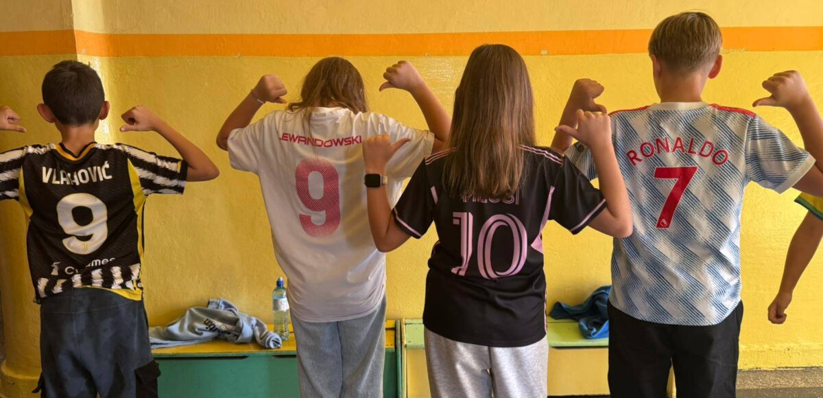 Uczniowie stoją plecami na szkolnym korytarzu, prezentując różne koszulki drużyn piłkarskich