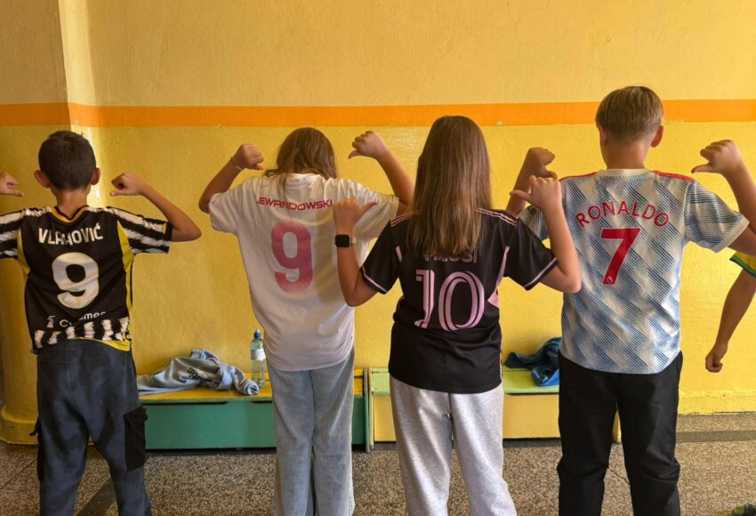 Uczniowie stoją plecami na szkolnym korytarzu, prezentując różne koszulki drużyn piłkarskich