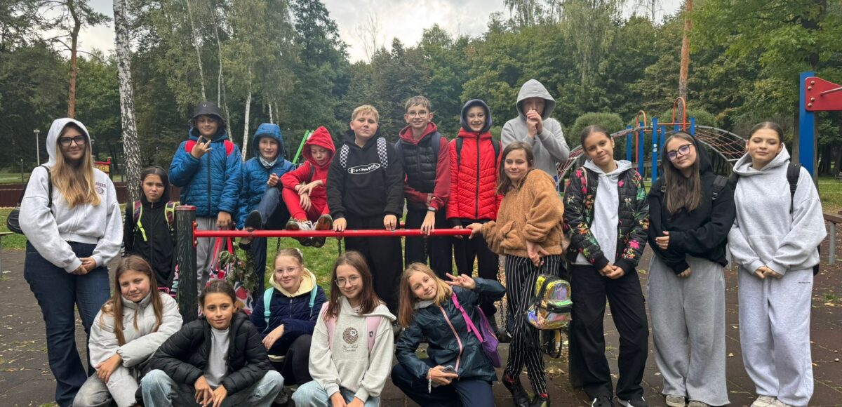 Grupa uczniów klasy VI pozująca wspólnie na szkolnym placu zabaw podczas wyjścia klasowego; dzieci stoją i siedzą przy drabinkach, uśmiechają się i mają na sobie bluzy oraz kurtki, w tle widać drzewa i park.