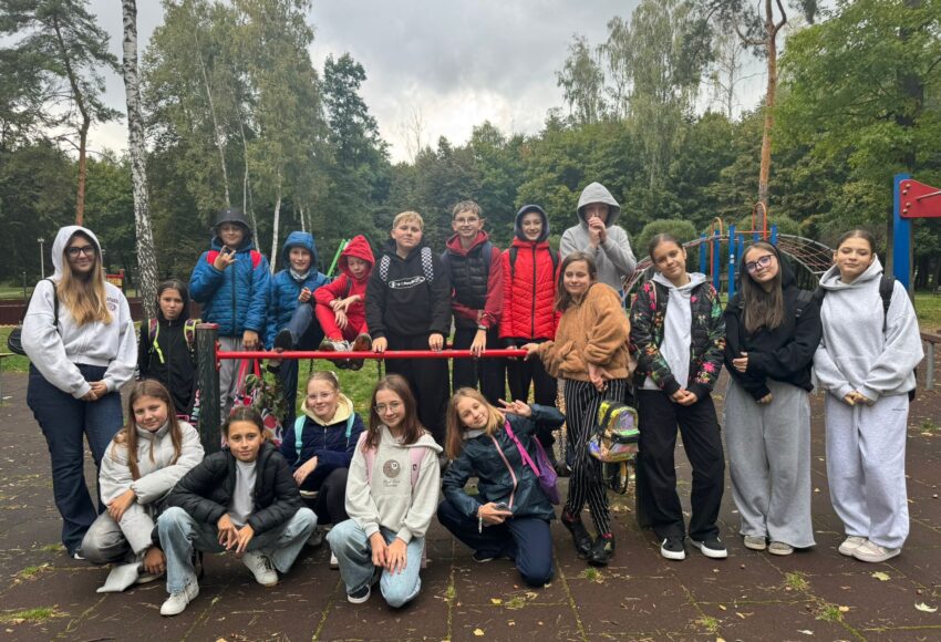 Grupa uczniów klasy VI pozująca wspólnie na szkolnym placu zabaw podczas wyjścia klasowego; dzieci stoją i siedzą przy drabinkach, uśmiechają się i mają na sobie bluzy oraz kurtki, w tle widać drzewa i park.