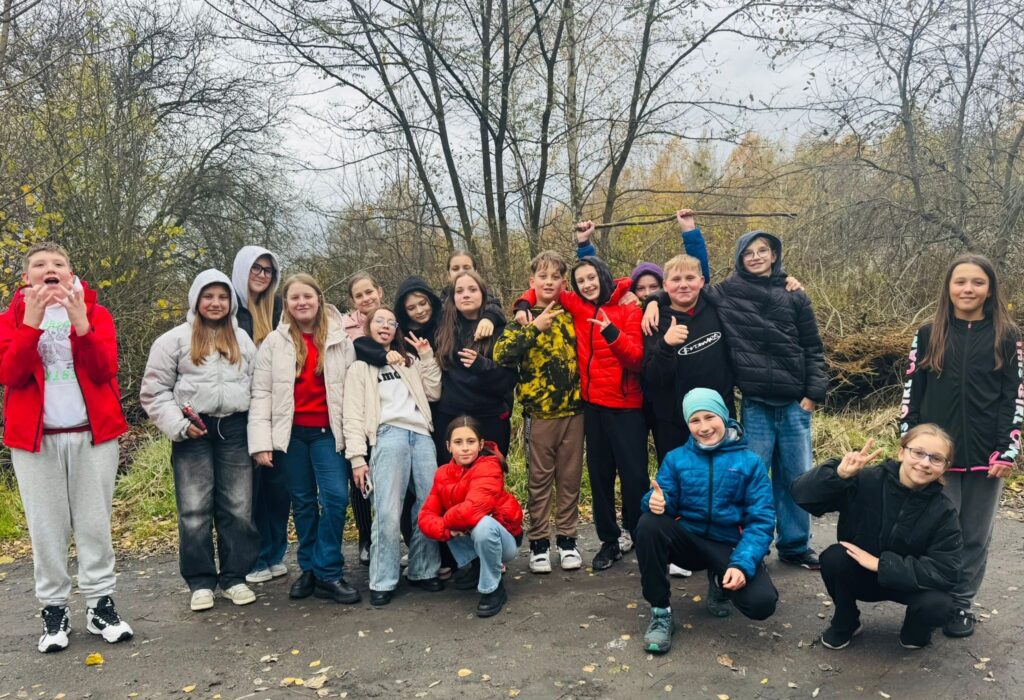 „Grupa uczniów klasy VI spacerująca ścieżką wśród jesiennej przyrody; uczniowie ubrani w kurtki, w tle drzewa z kolorowymi, jesiennymi liśćmi.”