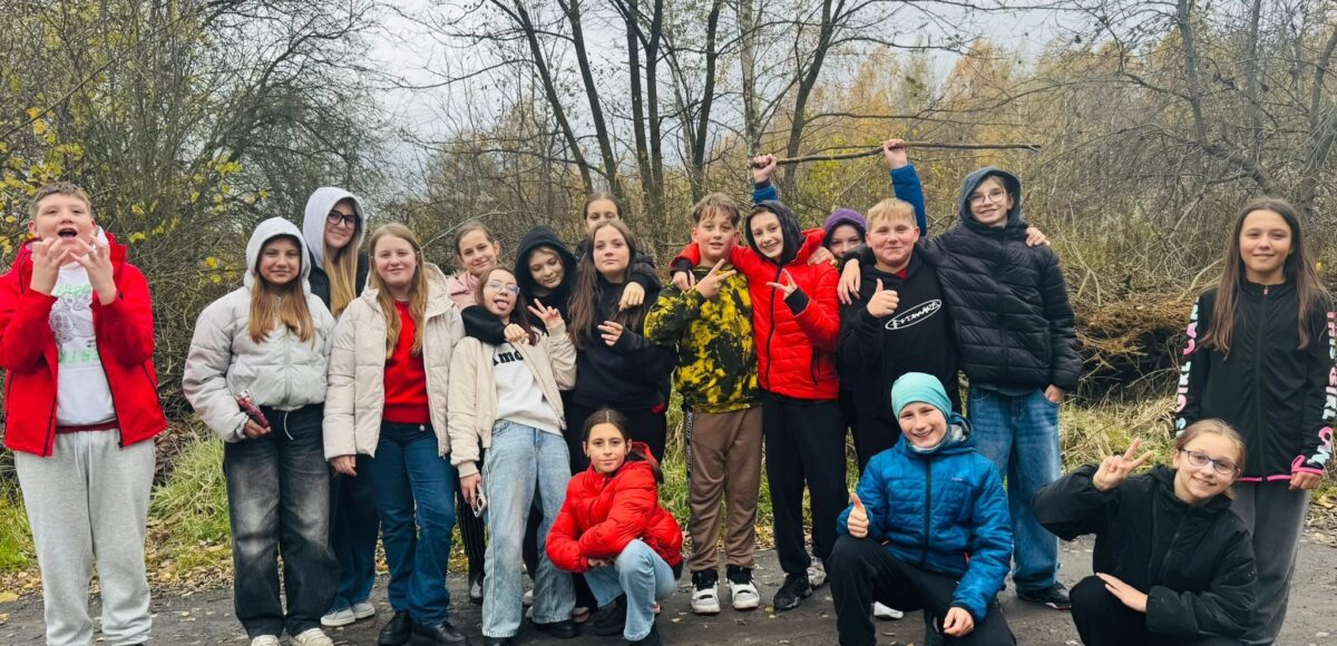 „Grupa uczniów klasy VI spacerująca ścieżką wśród jesiennej przyrody; uczniowie ubrani w kurtki, w tle drzewa z kolorowymi, jesiennymi liśćmi.”