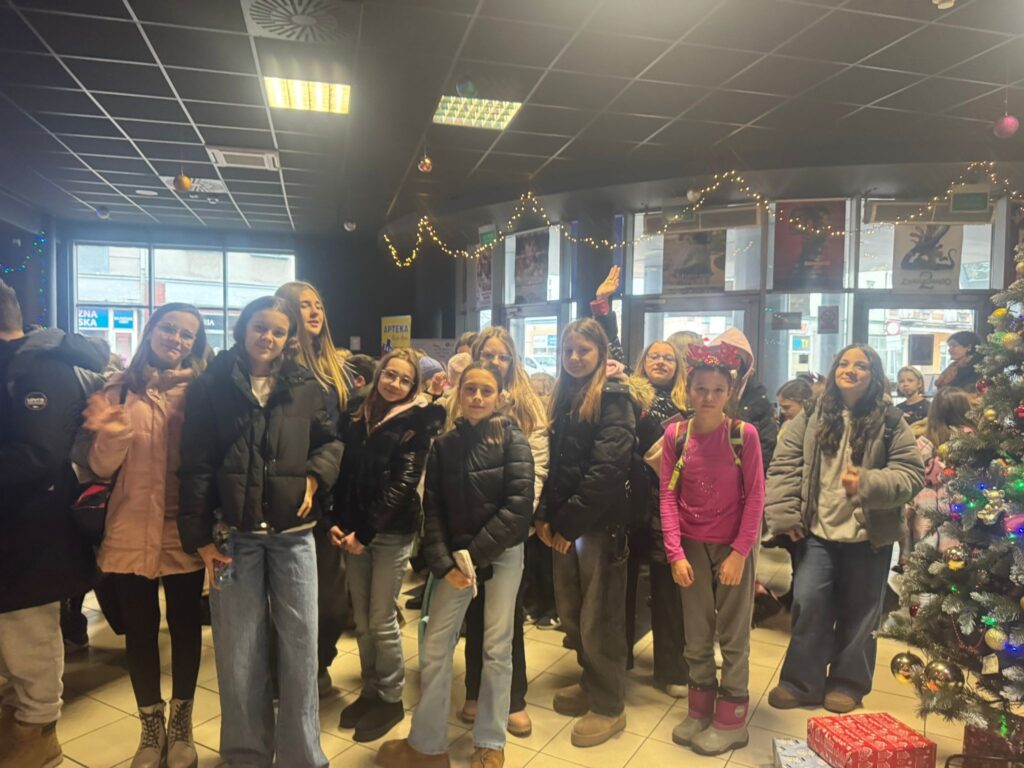 Grupa uczniów klasy VI stoi w holu kina udekorowanym świątecznymi lampkami i choinką. Uczniowie uśmiechają się i pozują do zdjęcia przed wejściem na seans mikołajkowy.