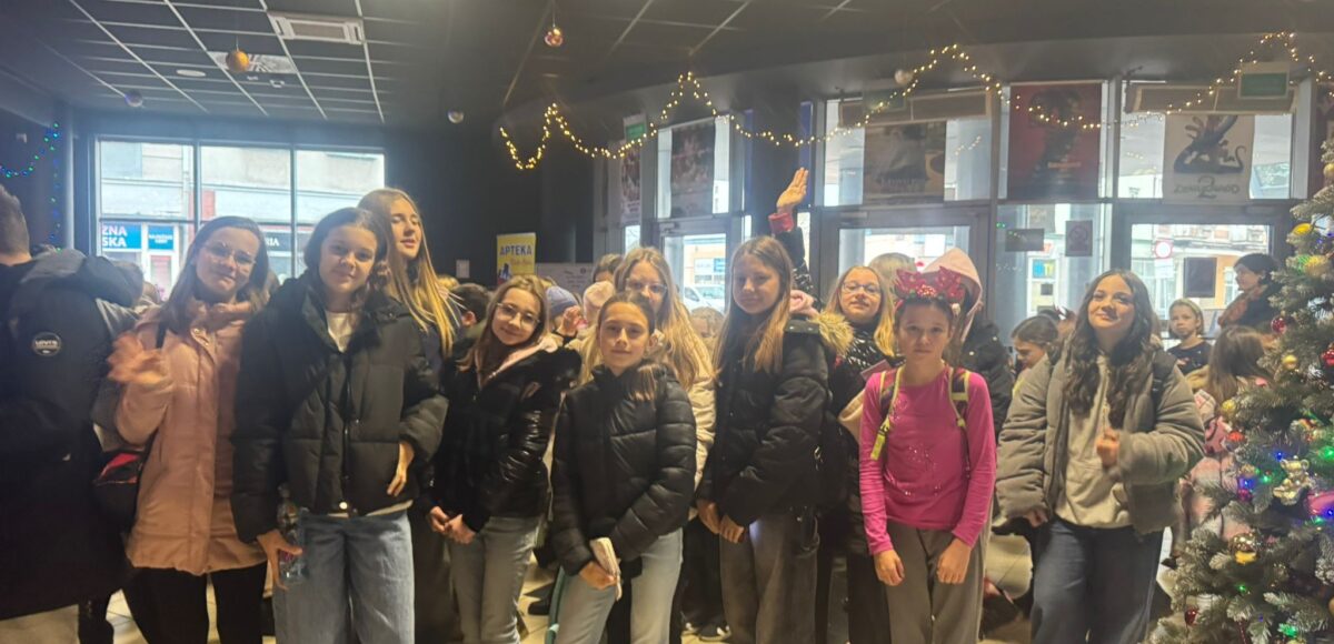 Grupa uczniów klasy VI stoi w holu kina udekorowanym świątecznymi lampkami i choinką. Uczniowie uśmiechają się i pozują do zdjęcia przed wejściem na seans mikołajkowy.