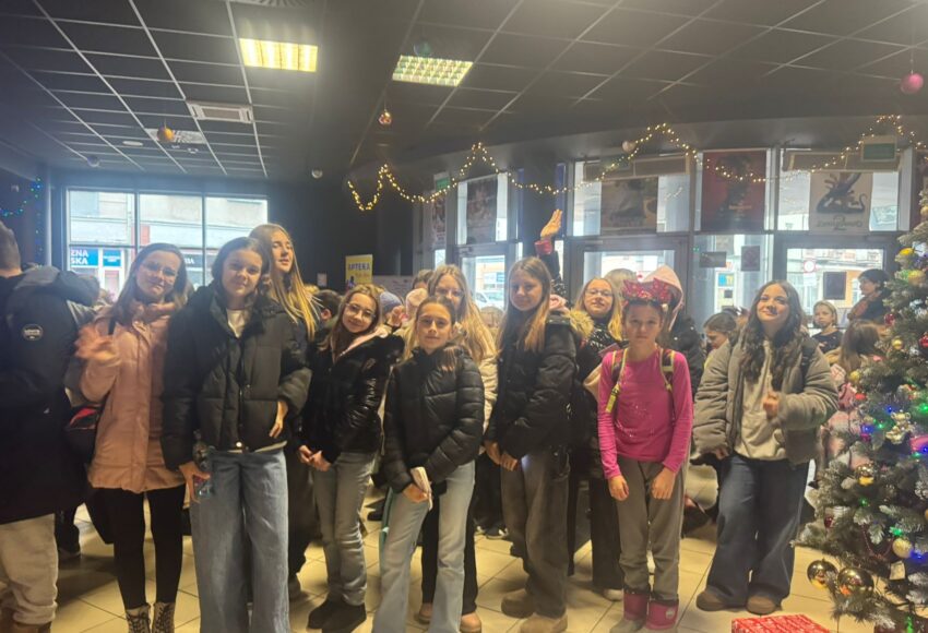 Grupa uczniów klasy VI stoi w holu kina udekorowanym świątecznymi lampkami i choinką. Uczniowie uśmiechają się i pozują do zdjęcia przed wejściem na seans mikołajkowy.