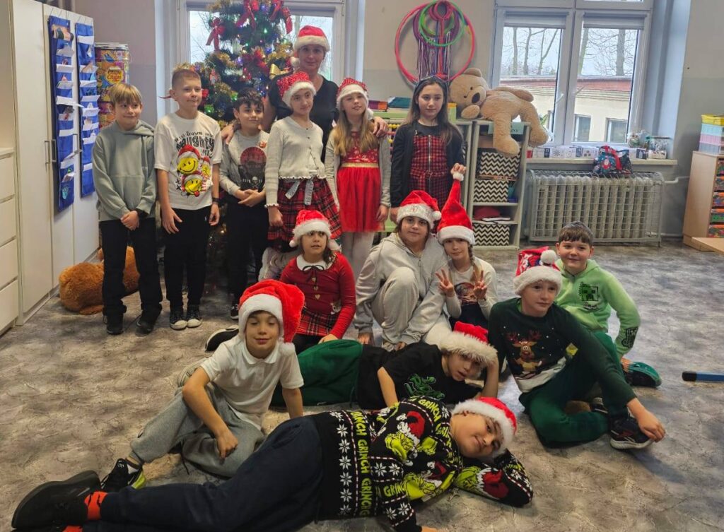 Zdjęcie przedstawia uczniów klasy 3 z wychowawczynią ubranych w czapki mikołajkowe na tle ubranej choinki