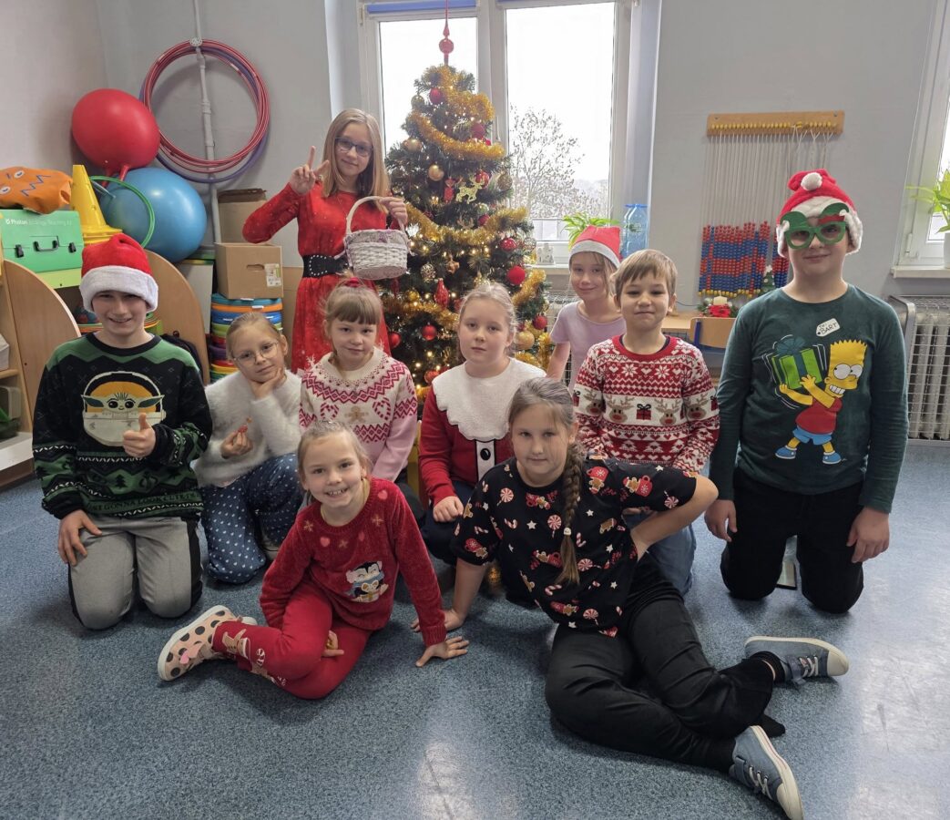 Grupa uczniów w świątecznych sweterkach pozująca przy udekorowanej choince podczas szkolnego Dnia Świątecznego Sweterka; dzieci uśmiechają się i mają na sobie różnorodne zimowe i świąteczne motywy.