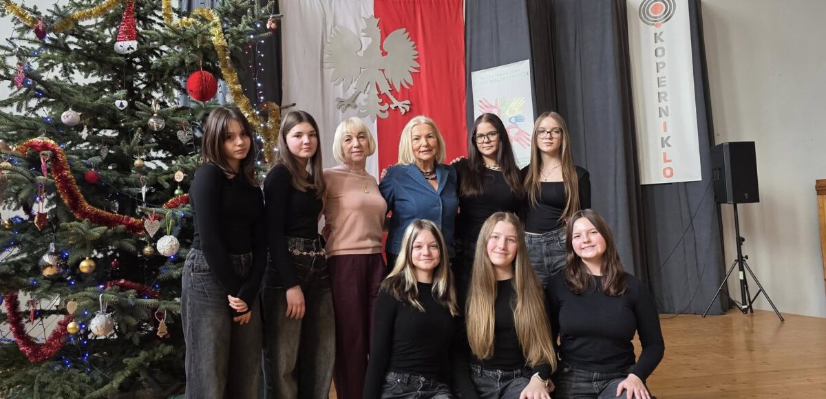 Zespół taneczny „Fantazja” wraz z opiekunką pozuje do zdjęcia na scenie I LO w Będzinie, obok udekorowanej świątecznej choinki i na tle godła oraz dekoracji wydarzenia.