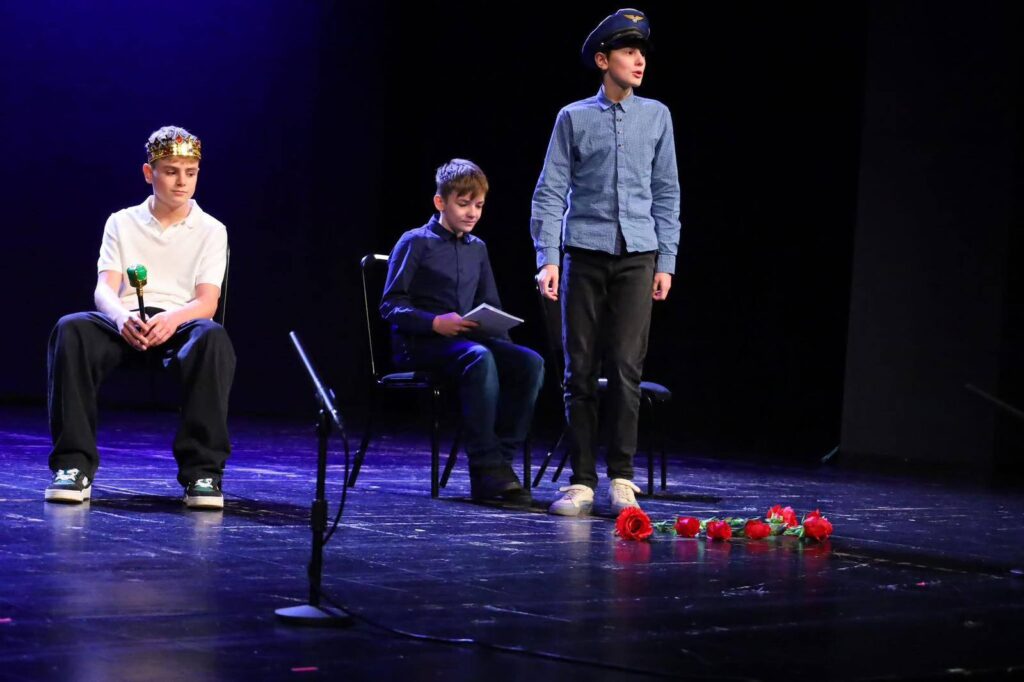 Uczniowie klasy 7a podczas występu teatralnego na scenie Pałacu Młodzieży w Katowicach; jeden z uczniów siedzi w koronie, drugi trzyma notes, a trzeci stoi w czapce pilota, na podłodze leżą czerwone róże.
