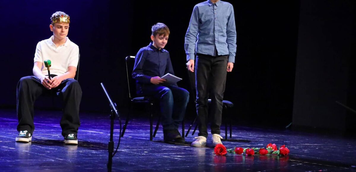 Uczniowie klasy 7a podczas występu teatralnego na scenie Pałacu Młodzieży w Katowicach; jeden z uczniów siedzi w koronie, drugi trzyma notes, a trzeci stoi w czapce pilota, na podłodze leżą czerwone róże.