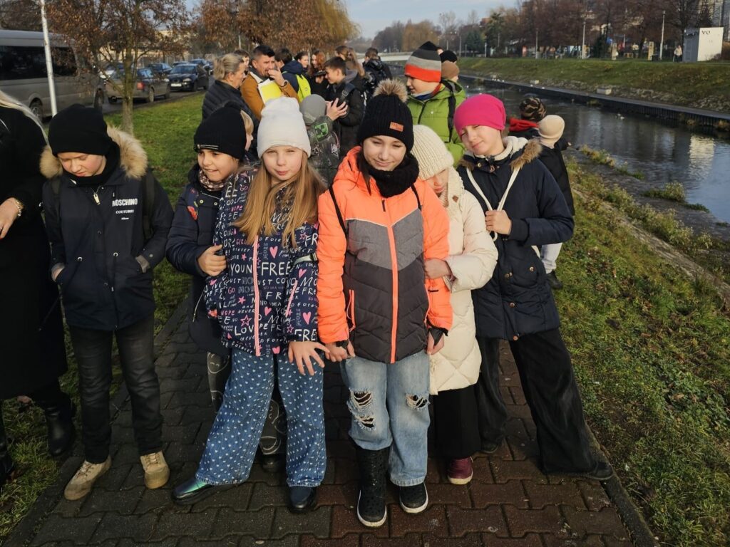 Uczniowie klasy trzeciej podczas spaceru terenowego nad Czarną Przemszą; grupa dzieci w zimowych kurtkach stoi na chodniku obok rzeki, uczestnicząc w warsztatach ekologicznych.