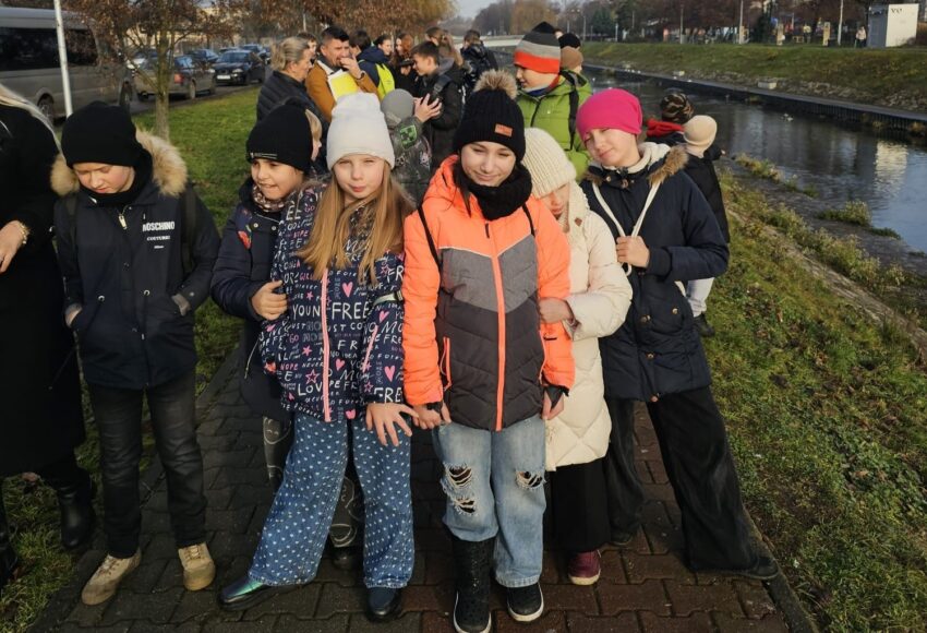 Uczniowie klasy trzeciej podczas spaceru terenowego nad Czarną Przemszą; grupa dzieci w zimowych kurtkach stoi na chodniku obok rzeki, uczestnicząc w warsztatach ekologicznych.