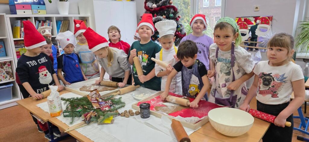 Uczniowie klasy 1 wspólnie wałkują ciasto na pierniki przy stołach w sali szkolnej. Dzieci mają czapki Mikołaja i fartuszki, a w tle widać udekorowaną choinkę oraz świąteczne ozdoby.