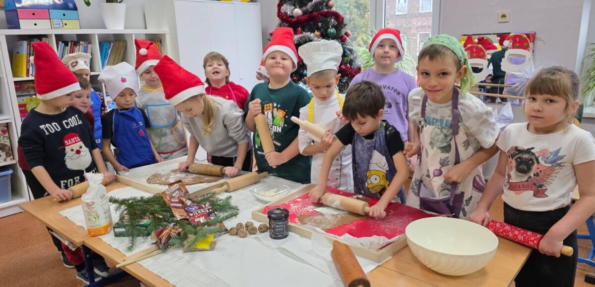 Uczniowie klasy 1 wspólnie wałkują ciasto na pierniki przy stołach w sali szkolnej. Dzieci mają czapki Mikołaja i fartuszki, a w tle widać udekorowaną choinkę oraz świąteczne ozdoby.