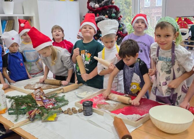 Uczniowie klasy 1 wspólnie wałkują ciasto na pierniki przy stołach w sali szkolnej. Dzieci mają czapki Mikołaja i fartuszki, a w tle widać udekorowaną choinkę oraz świąteczne ozdoby.