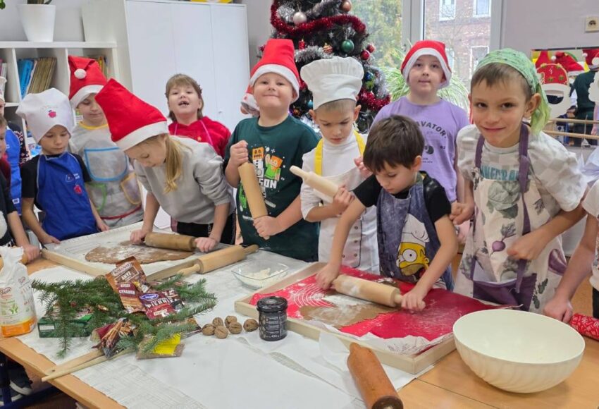 Uczniowie klasy 1 wspólnie wałkują ciasto na pierniki przy stołach w sali szkolnej. Dzieci mają czapki Mikołaja i fartuszki, a w tle widać udekorowaną choinkę oraz świąteczne ozdoby.