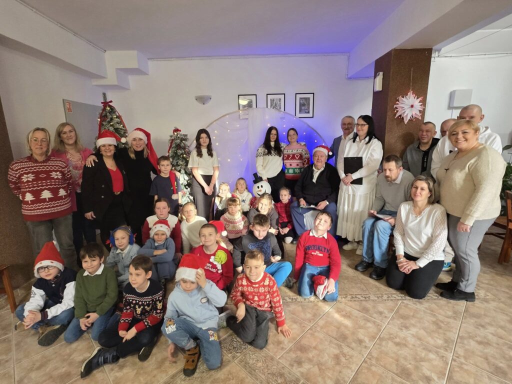 Uczniowie oraz nauczyciele Szkoły Podstawowej nr 6 podczas świątecznej wizyty w Domu Pomocy Społecznej w Będzinie-Grodźcu. Wspólne zdjęcie dzieci, seniorów i opiekunów na tle świątecznych dekoracji, choinek i kolędowej scenografii.