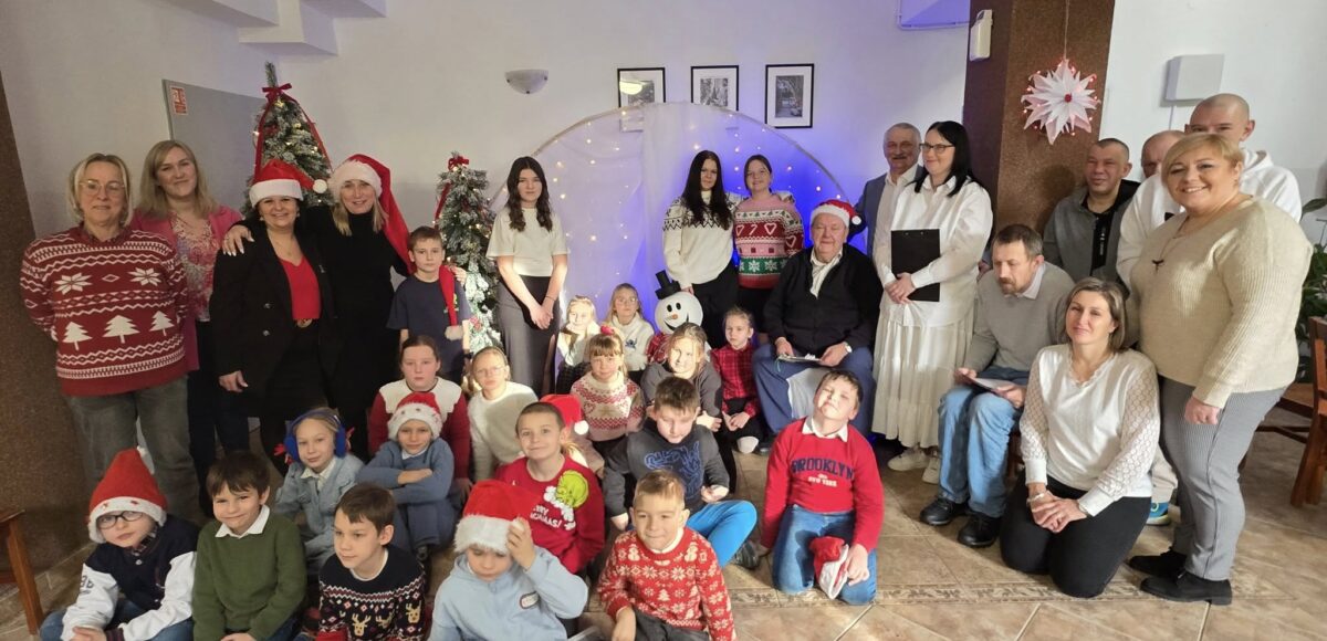 Uczniowie oraz nauczyciele Szkoły Podstawowej nr 6 podczas świątecznej wizyty w Domu Pomocy Społecznej w Będzinie-Grodźcu. Wspólne zdjęcie dzieci, seniorów i opiekunów na tle świątecznych dekoracji, choinek i kolędowej scenografii.