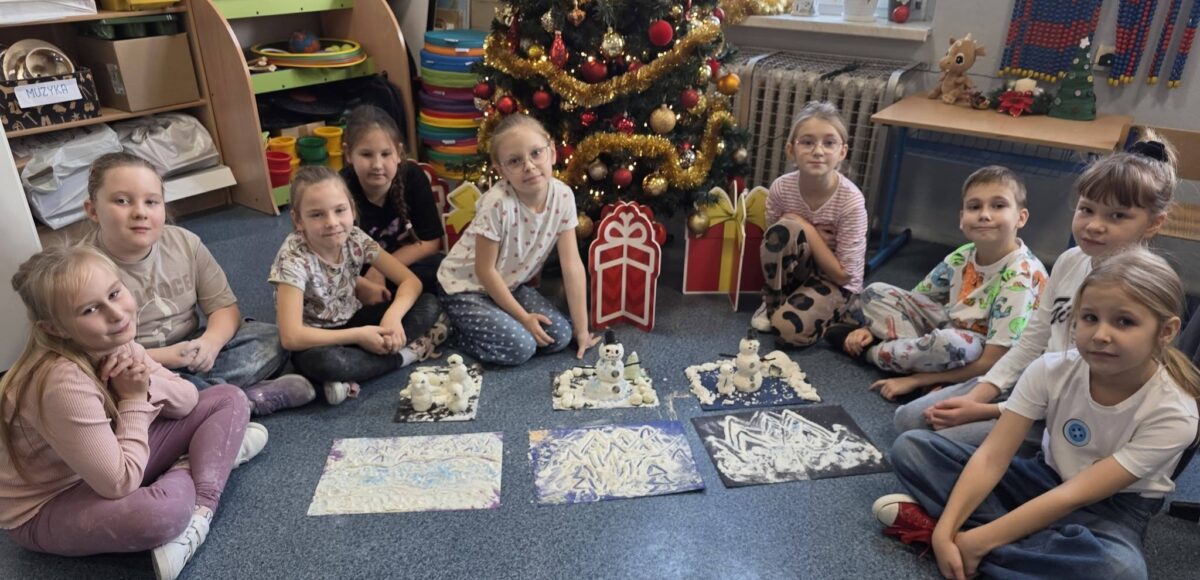 Uczniowie klasy 2a siedzą w kole na dywanie przy udekorowanej choince. Przed nimi znajdują się prace plastyczne przedstawiające zimowe krajobrazy i bałwanki wykonane w ramach projektu „Zabawa sztuką”.