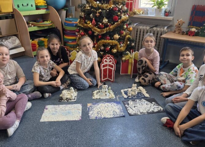 Uczniowie klasy 2a siedzą w kole na dywanie przy udekorowanej choince. Przed nimi znajdują się prace plastyczne przedstawiające zimowe krajobrazy i bałwanki wykonane w ramach projektu „Zabawa sztuką”.