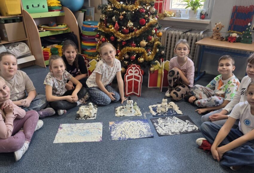 Uczniowie klasy 2a siedzą w kole na dywanie przy udekorowanej choince. Przed nimi znajdują się prace plastyczne przedstawiające zimowe krajobrazy i bałwanki wykonane w ramach projektu „Zabawa sztuką”.