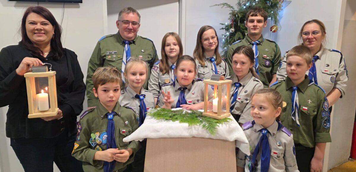 Przekazanie Betlejemskiego Światła Pokoju – zuchy i harcerze w mundurach stoją razem z Wicedyrektor Szkoły, prezentując zapalone lampiony z Betlejemskim Światłem Pokoju w szkolnej sali, na tle świątecznej dekoracji.