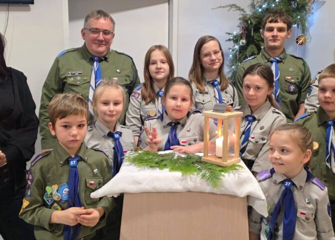 Przekazanie Betlejemskiego Światła Pokoju – zuchy i harcerze w mundurach stoją razem z Wicedyrektor Szkoły, prezentując zapalone lampiony z Betlejemskim Światłem Pokoju w szkolnej sali, na tle świątecznej dekoracji.
