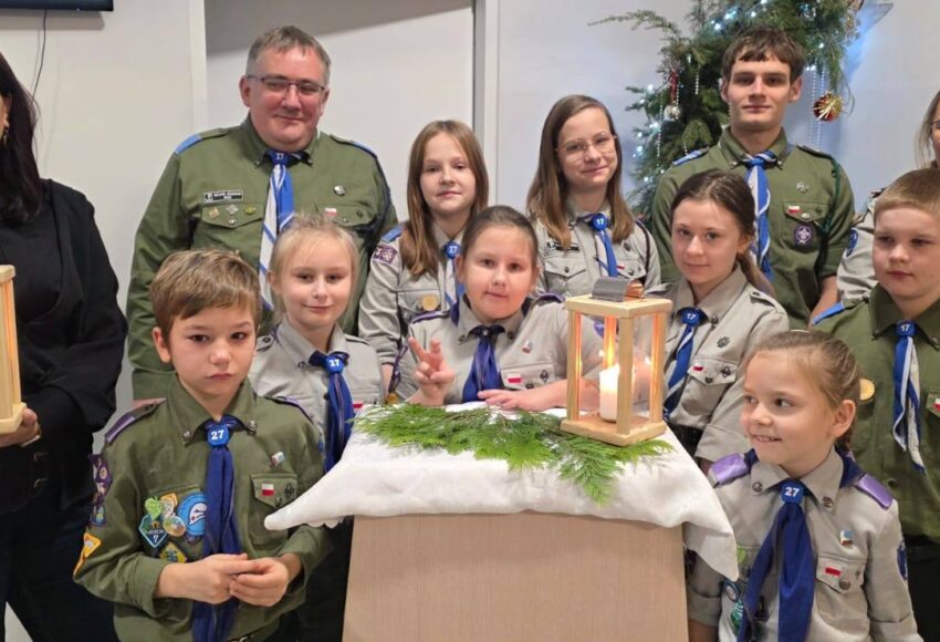Przekazanie Betlejemskiego Światła Pokoju – zuchy i harcerze w mundurach stoją razem z Wicedyrektor Szkoły, prezentując zapalone lampiony z Betlejemskim Światłem Pokoju w szkolnej sali, na tle świątecznej dekoracji.