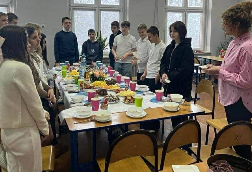 Uczniowie zgromadzeni przy wspólnym stole podczas klasowej wigilii. Na stole znajdują się świąteczne potrawy, a nauczycielka prowadzi spotkanie w sali lekcyjnej.