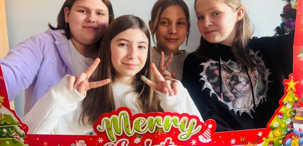 Cztery uśmiechnięte uczennice pozują do zdjęcia w świątecznej ramce z napisem „Merry Christmas”, trzymając palce w geście pokoju. W tle widoczne są świąteczne dekoracje.