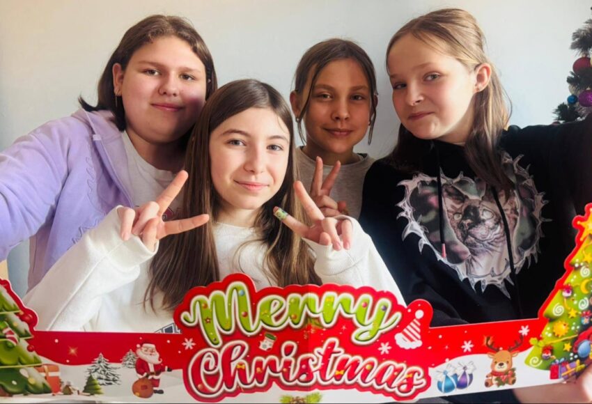 Cztery uśmiechnięte uczennice pozują do zdjęcia w świątecznej ramce z napisem „Merry Christmas”, trzymając palce w geście pokoju. W tle widoczne są świąteczne dekoracje.