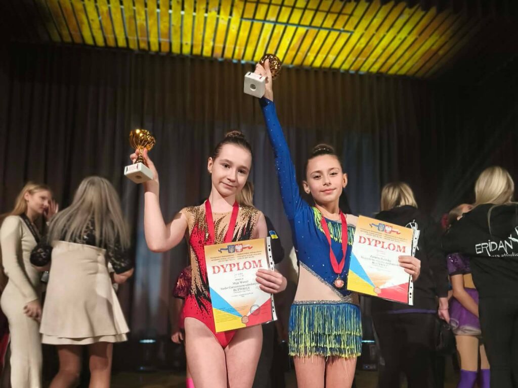 Uczennice Szkoły Podstawowej nr 6 prezentujące dyplomy i puchary podczas konkursu tanecznego „Polish Art Wave”. Dziewczęta stoją na scenie w strojach tanecznych i unoszą nagrody w górze.