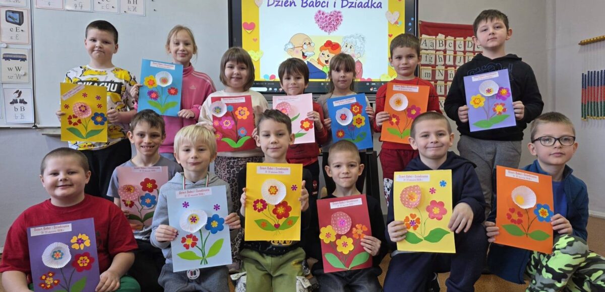 „Uczniowie klasy I prezentują własnoręcznie wykonane laurki z okazji Dnia Babci i Dziadka; dzieci trzymają kolorowe prace plastyczne w sali lekcyjnej, w tle widoczna tablica z napisem »Dzień Babci i Dziadka