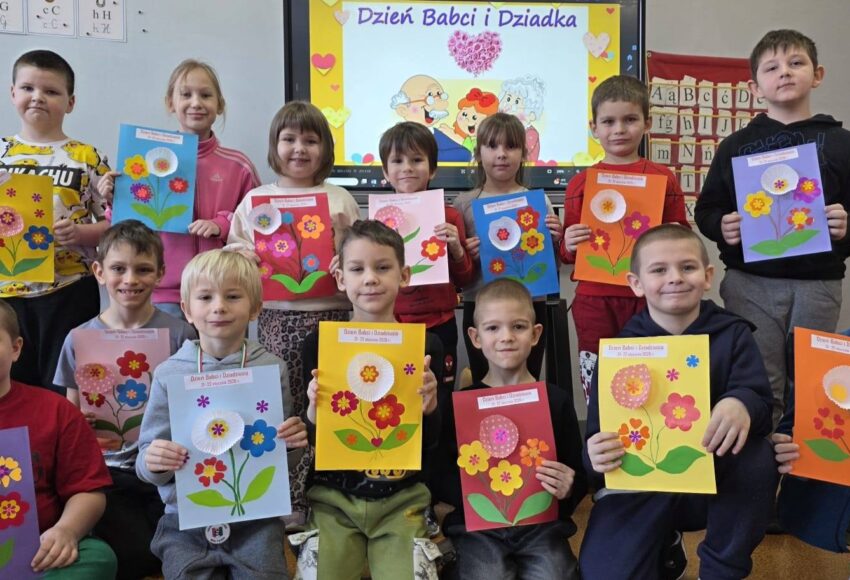 „Uczniowie klasy I prezentują własnoręcznie wykonane laurki z okazji Dnia Babci i Dziadka; dzieci trzymają kolorowe prace plastyczne w sali lekcyjnej, w tle widoczna tablica z napisem »Dzień Babci i Dziadka