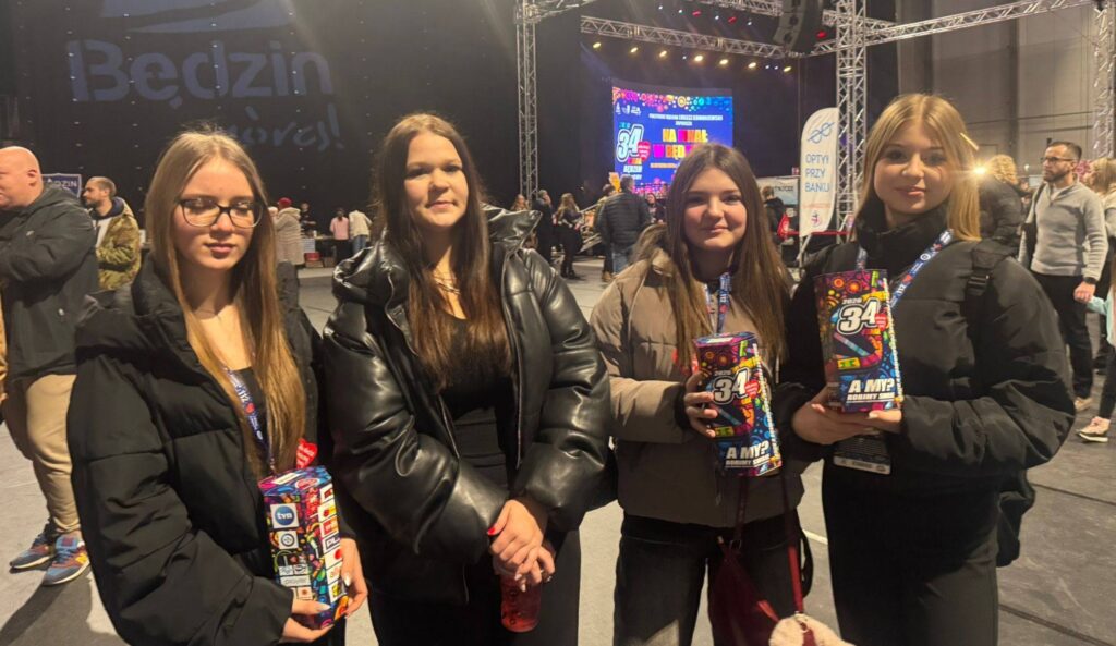 Szkolne wolontariuszki podczas 34. Finału Wielkiej Orkiestry Świątecznej Pomocy w hali Będzin Arena, trzymające puszki WOŚP na tle sceny wydarzenia.