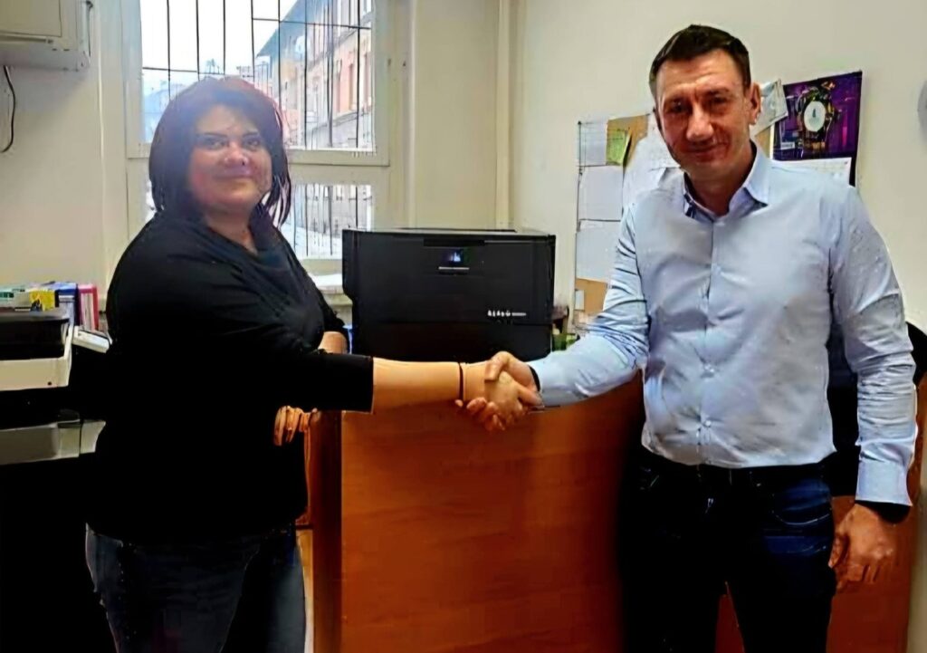 Przedstawiciel firmy Konica Minolta przekazuje nowoczesną drukarkę biurową przedstawicielce szkoły – uścisk dłoni na tle nowego urządzenia w pomieszczeniu biurowym.