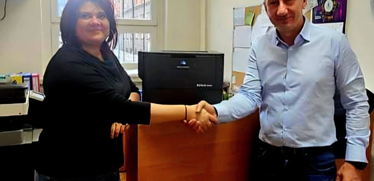 Przedstawiciel firmy Konica Minolta przekazuje nowoczesną drukarkę biurową przedstawicielce szkoły – uścisk dłoni na tle nowego urządzenia w pomieszczeniu biurowym.