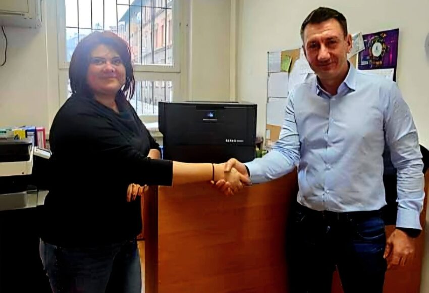 Przedstawiciel firmy Konica Minolta przekazuje nowoczesną drukarkę biurową przedstawicielce szkoły – uścisk dłoni na tle nowego urządzenia w pomieszczeniu biurowym.