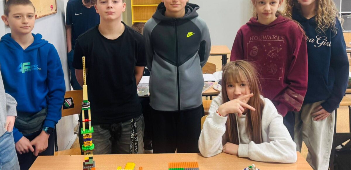 Uczniowie stoją przy ławkach w sali lekcyjnej, prezentując zbudowane z klocków LEGO konstrukcje podczas szkolnego LEGO Challenge.