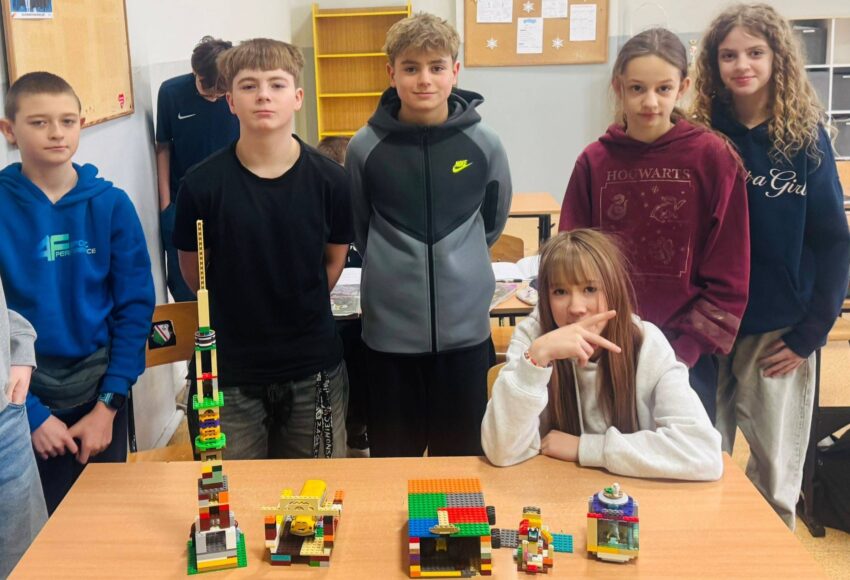 Uczniowie stoją przy ławkach w sali lekcyjnej, prezentując zbudowane z klocków LEGO konstrukcje podczas szkolnego LEGO Challenge.
