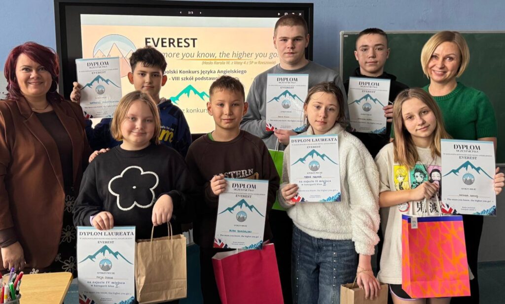 Uczniowie wraz z nauczycielami prezentują dyplomy i nagrody zdobyte w Ogólnopolskim Konkursie Języka Angielskiego EVEREST w szkolnej sali lekcyjnej.