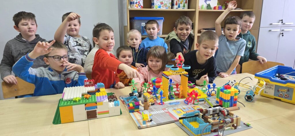 „Uczniowie klasy I podczas zajęć w pracowni Laboratorium Przyszłości budują konstrukcje z klocków na wspólnym stole, pracując zespołowo nad kolorowymi projektami.”