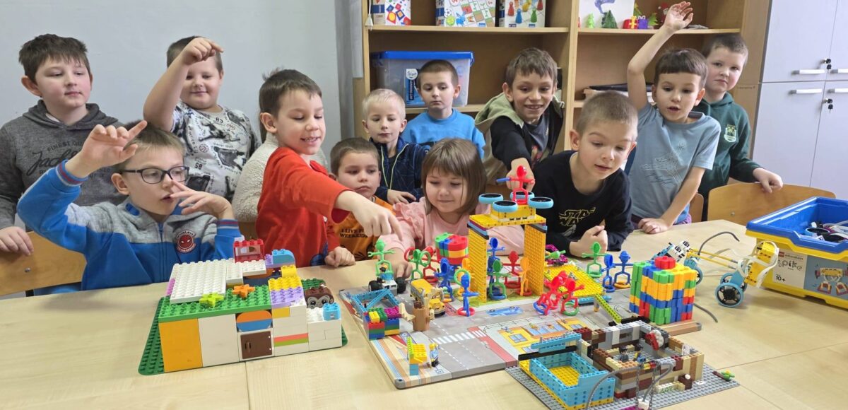 „Uczniowie klasy I podczas zajęć w pracowni Laboratorium Przyszłości budują konstrukcje z klocków na wspólnym stole, pracując zespołowo nad kolorowymi projektami.”