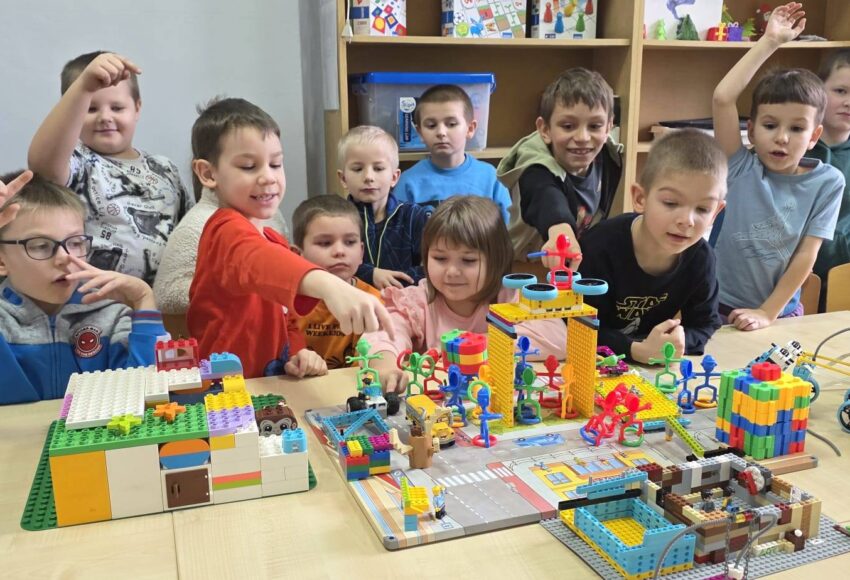 „Uczniowie klasy I podczas zajęć w pracowni Laboratorium Przyszłości budują konstrukcje z klocków na wspólnym stole, pracując zespołowo nad kolorowymi projektami.”