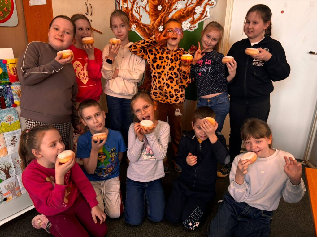 Uczniowie klasy II podczas obchodów Tłustego Czwartku w sali lekcyjnej, trzymający pączki i wspólnie świętujący tradycyjny dzień.