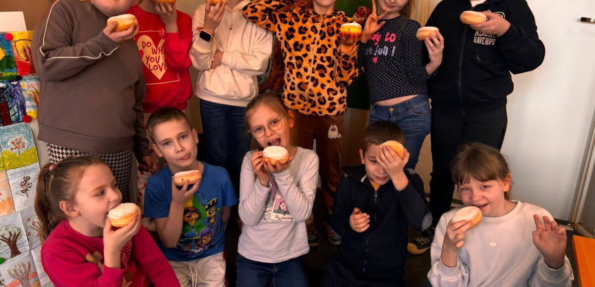 Uczniowie klasy II podczas obchodów Tłustego Czwartku w sali lekcyjnej, trzymający pączki i wspólnie świętujący tradycyjny dzień.