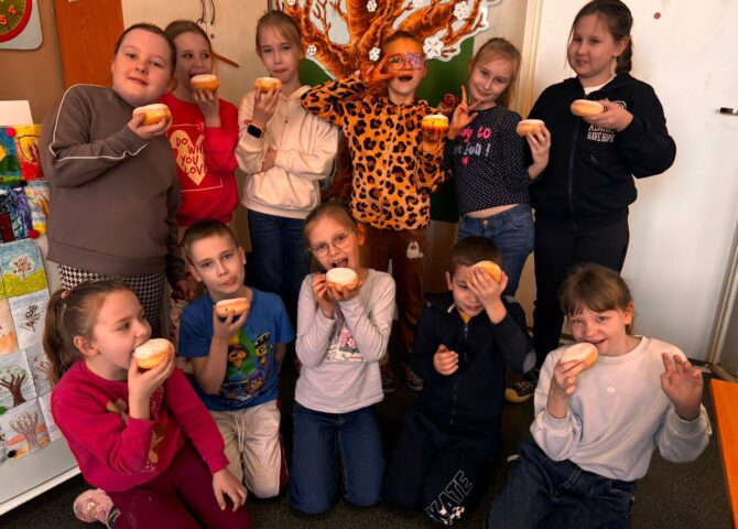 Uczniowie klasy II podczas obchodów Tłustego Czwartku w sali lekcyjnej, trzymający pączki i wspólnie świętujący tradycyjny dzień.