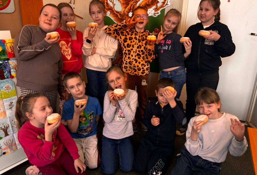 Uczniowie klasy II podczas obchodów Tłustego Czwartku w sali lekcyjnej, trzymający pączki i wspólnie świętujący tradycyjny dzień.