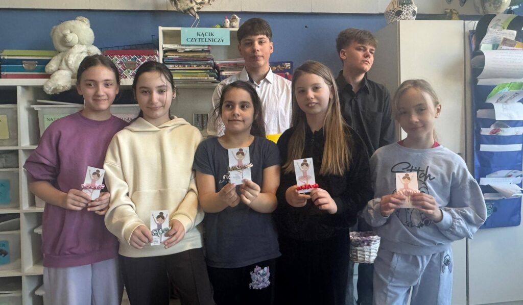 Uczniowie stoją w szkolnej sali przy kąciku czytelniczym, prezentując własnoręcznie przygotowane upominki z okazji Dnia Kobiet.